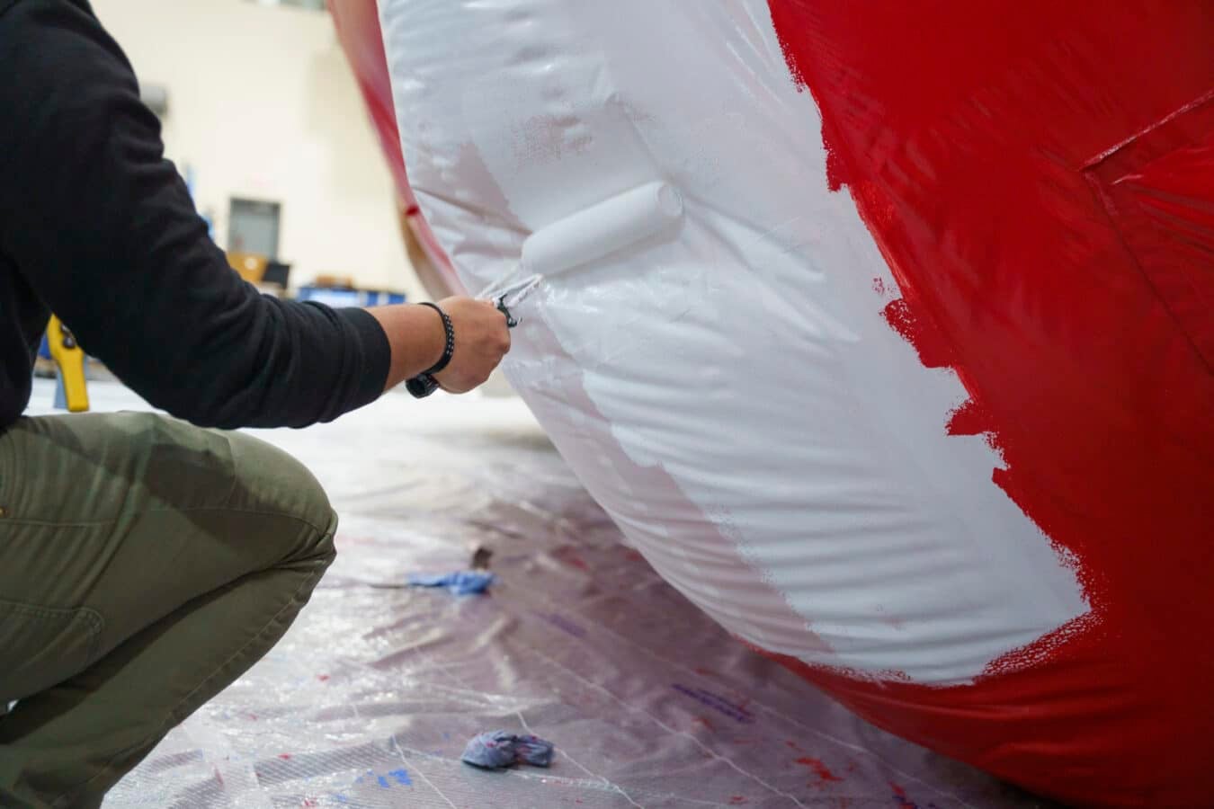 Person kneeling and rolling white paint onto a large surface with bright red paint on the edge nearby in a workshop setting