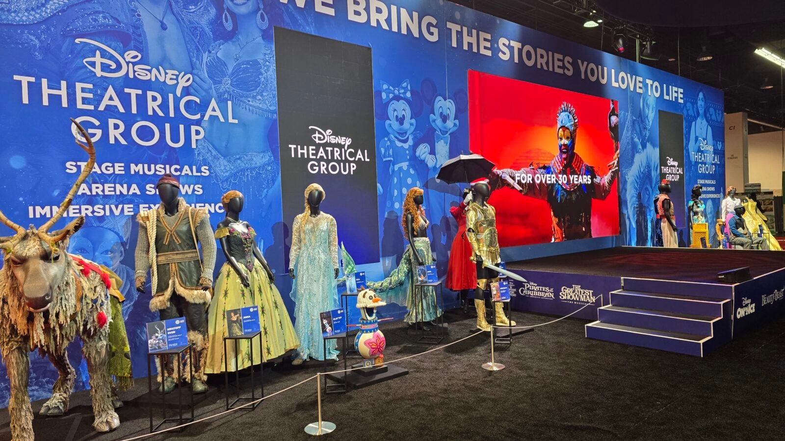 Display of Disney Theatrical Group costumes on mannequins against a blue promotional wall with banners and a red stage screen includes deer figure on left