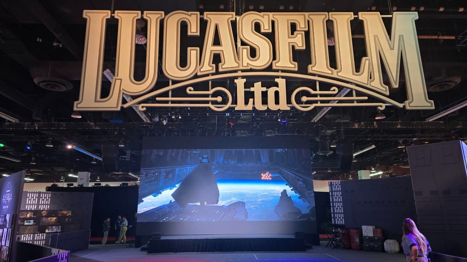 Large decorative sign reading LUCASFILM ltd hangs over a dark convention hall stage with a high ceiling and equipment above