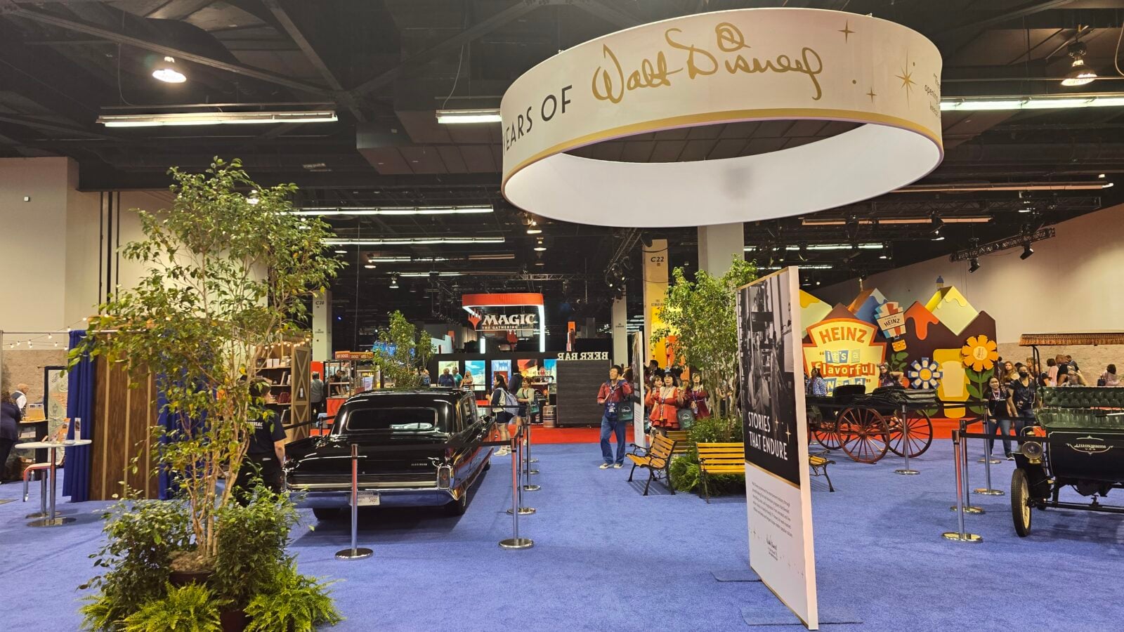Exhibition hall with a vintage car on a blue carpet surrounded by plants and colorful booths under a large circular hanging banner
