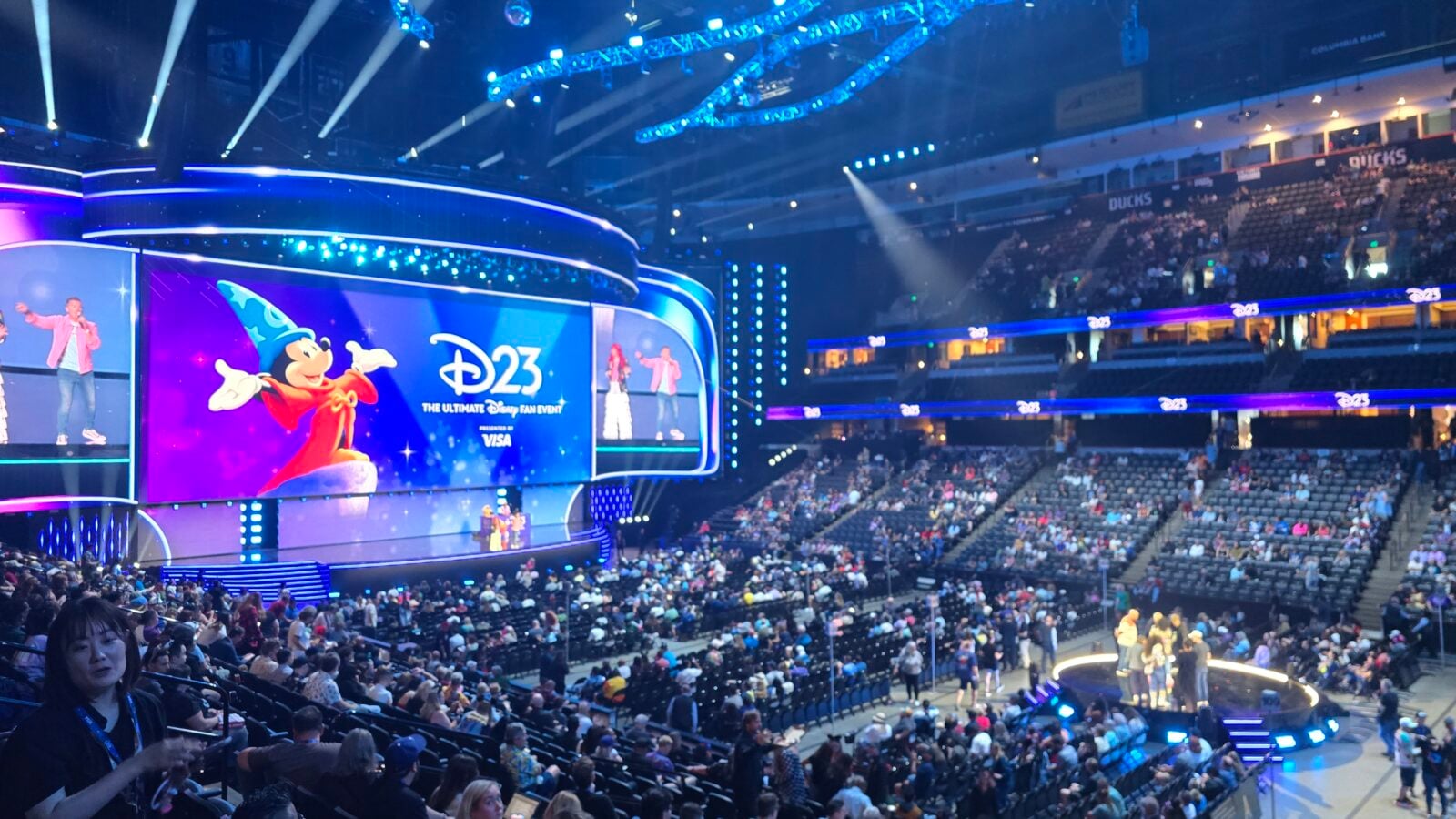 Stage at a D23 Disney fan event with Mickey Mouse on a large screen and a waving crowd in a lit arena