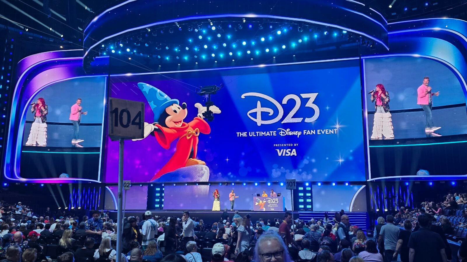Colorful D23 stage with Mickey Mouse in wizard hat on a large screen presenters on each side and a cheering crowd in front of a curved illuminated set