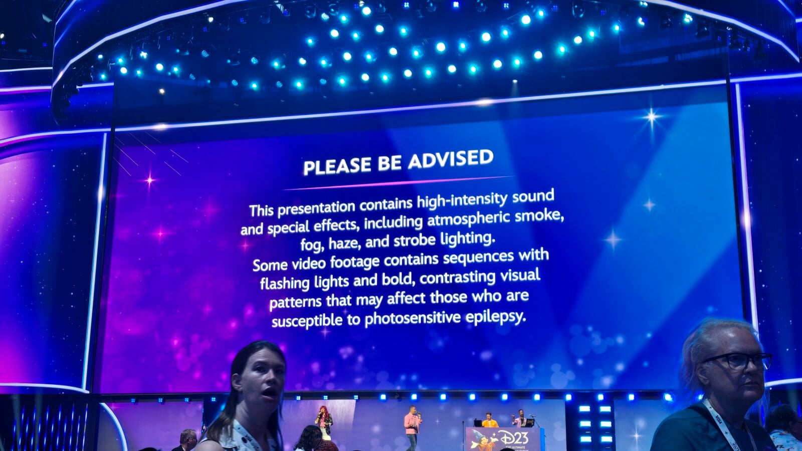 Stage with a large bluepurple screen showing the warning text PLEASE BE ADVISED about highintensity sound and flashing lights during the presentation performers visible on the lower stage