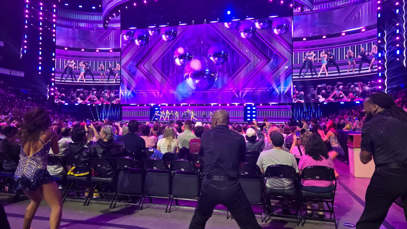 Large concert stage with purple lighting dancers performing on an elevated platform and a cheering audience in foreground