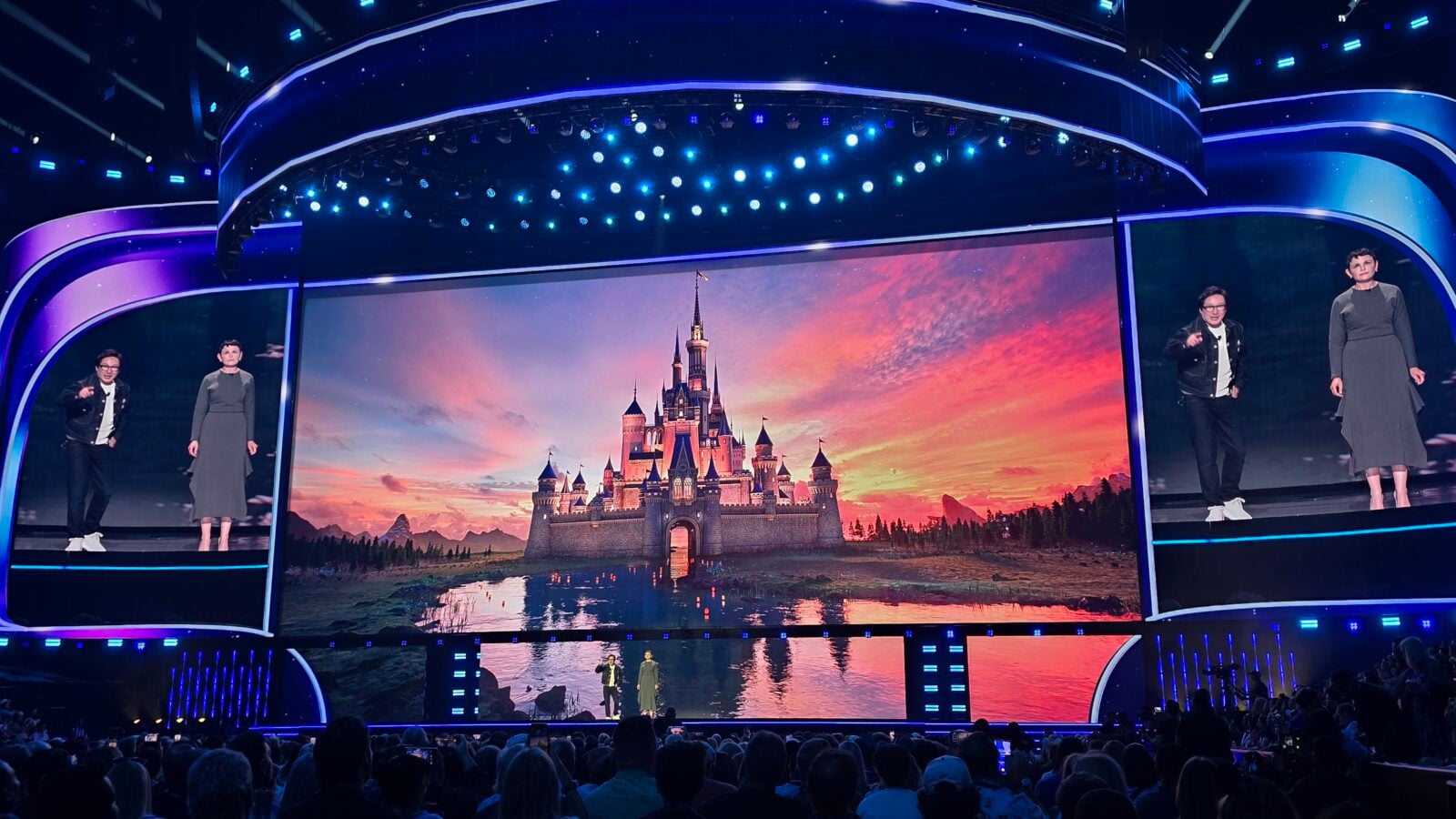 Stage show with a large screen showing a sunset castle performers on both sides and blue curved lighting across the set