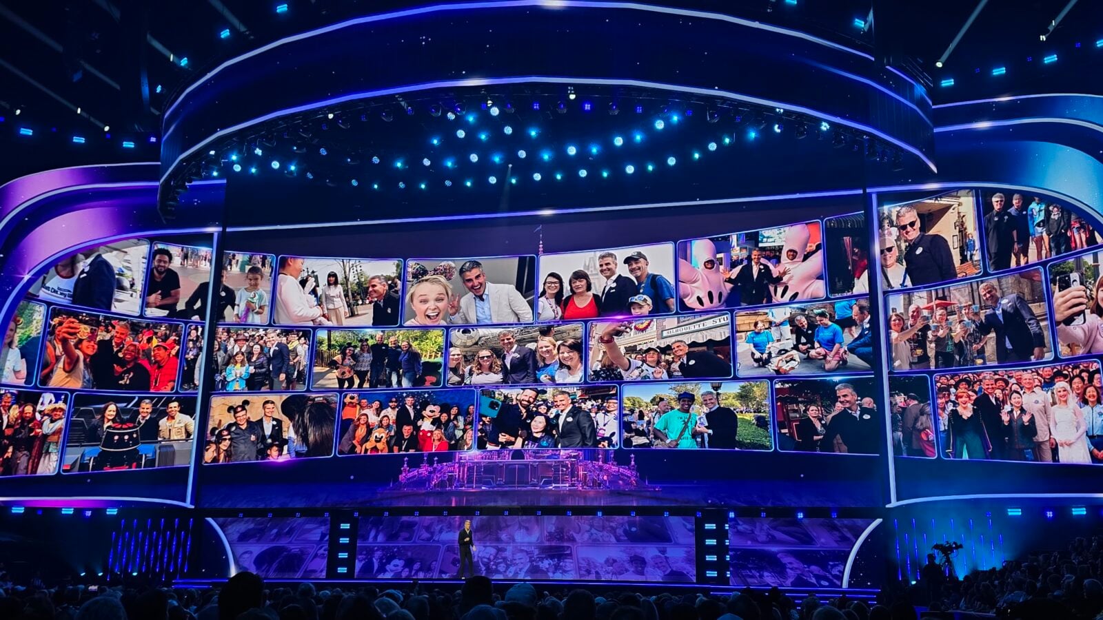 A large stage with a curved illuminated mosaic of photos showing diverse people while a presenter stands at the front under blue lighting