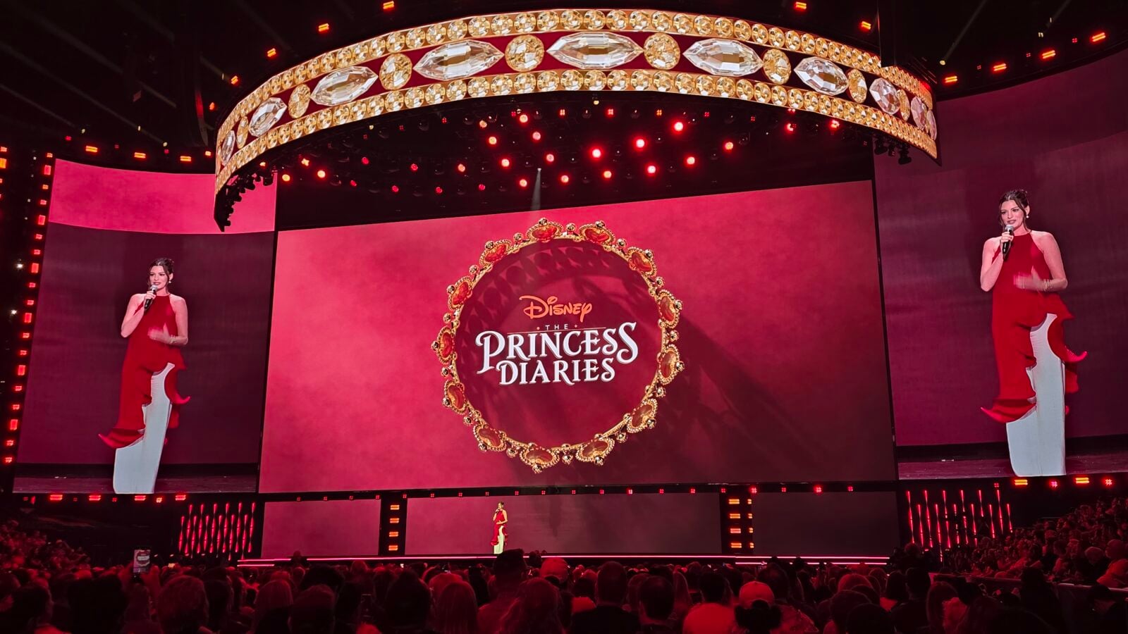 Stage event for Disneys Princess Diaries with two hosts in red dresses on either side of a large screen displaying a circular gold emblem and the title Disney The Princess Diaries above a red backdrop audience in foreground