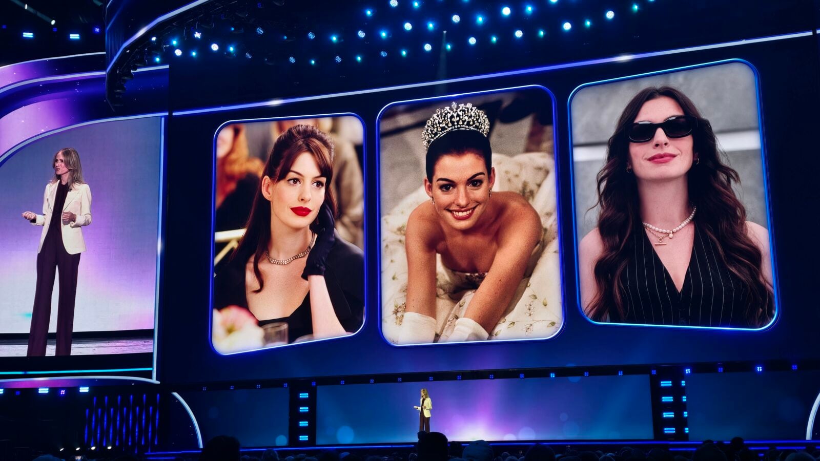 Stage with a presenter on a lit runway four tall blue framed screens show portraits of women wearing jewelry and crowns on display