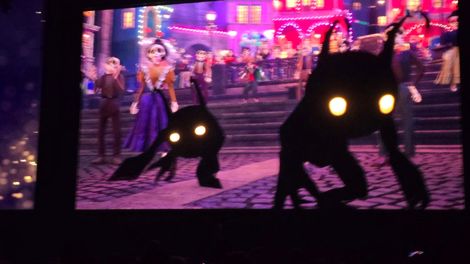 Stage performance with dancers in skeleton costumes under pink and purple lights dark foreground silhouettes with glowing yellow eyes