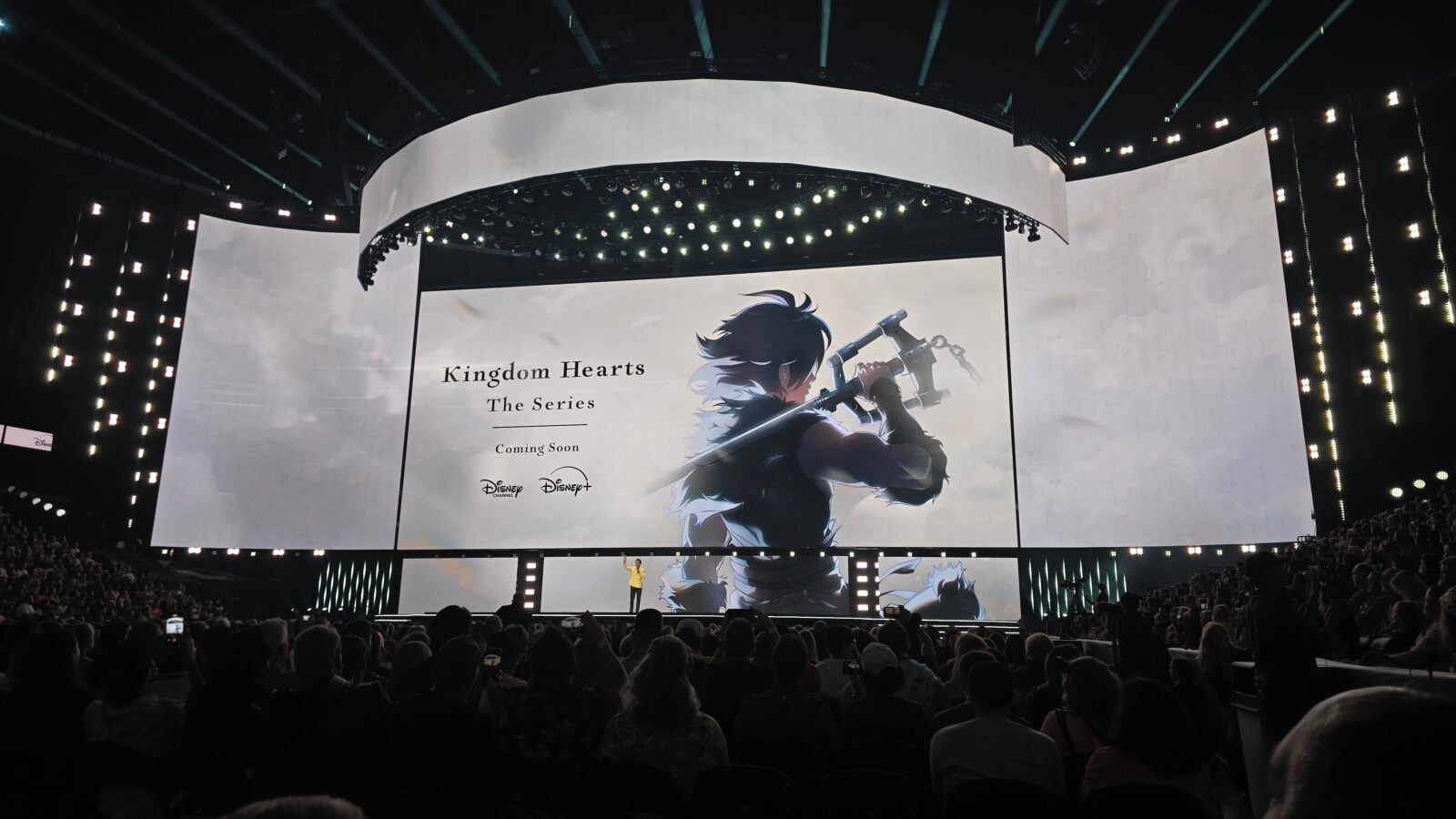 Stage reveal showing'Kingdom Hearts The Series Coming Soon' with a warrior illustration on a large screen and audience in front of the stage.