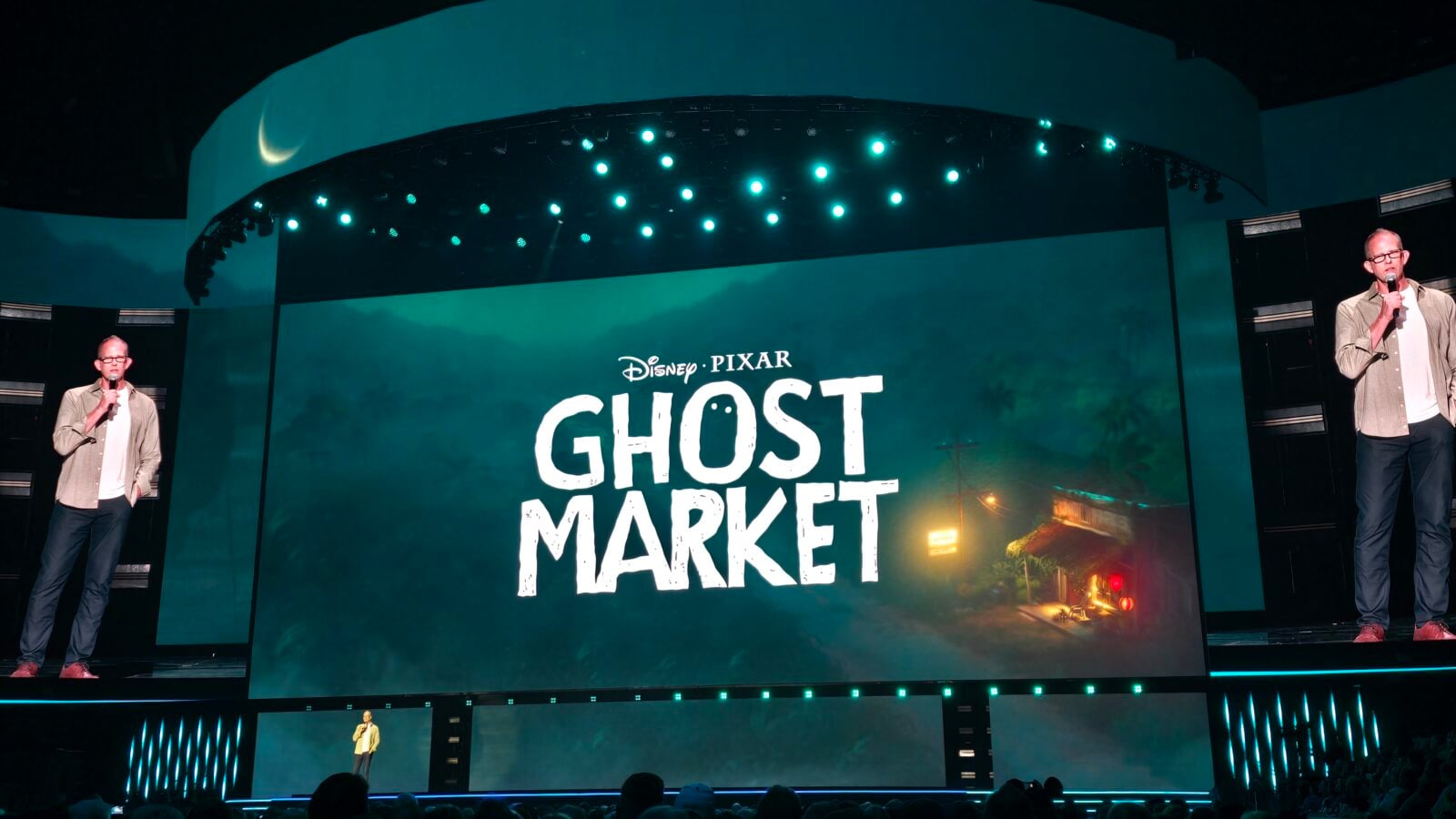 Two presenters on a dark stage with microphones a large screen behind them reads Disney Pixar Ghost Market in teal lighting