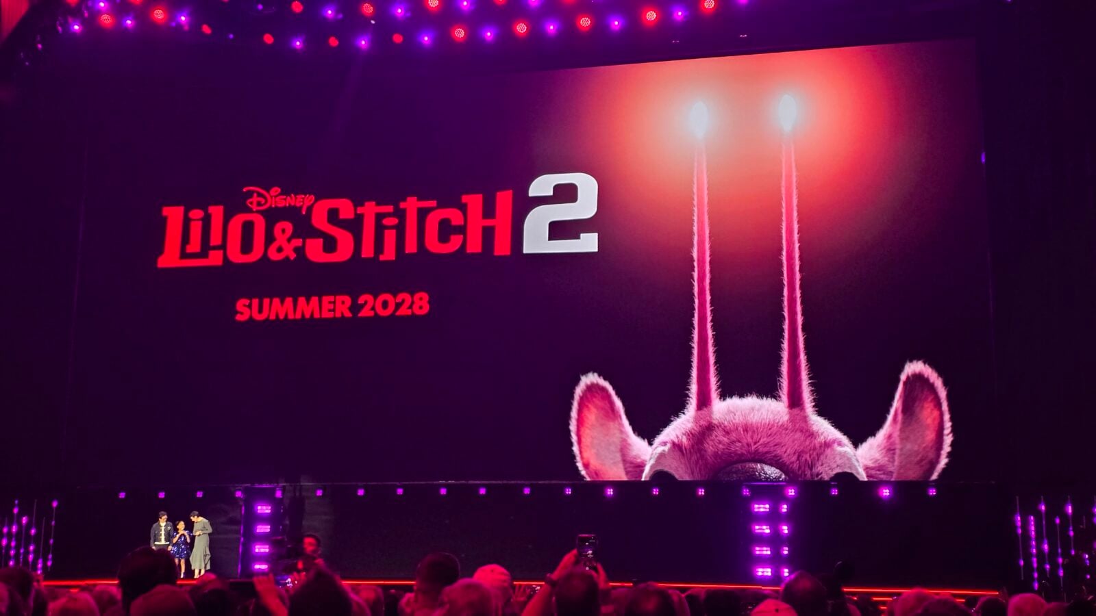 Stage with a large screen showing Lilo  Stitch 2 and Summer 2028 in red letters purple pink lights and an audience in the foreground
