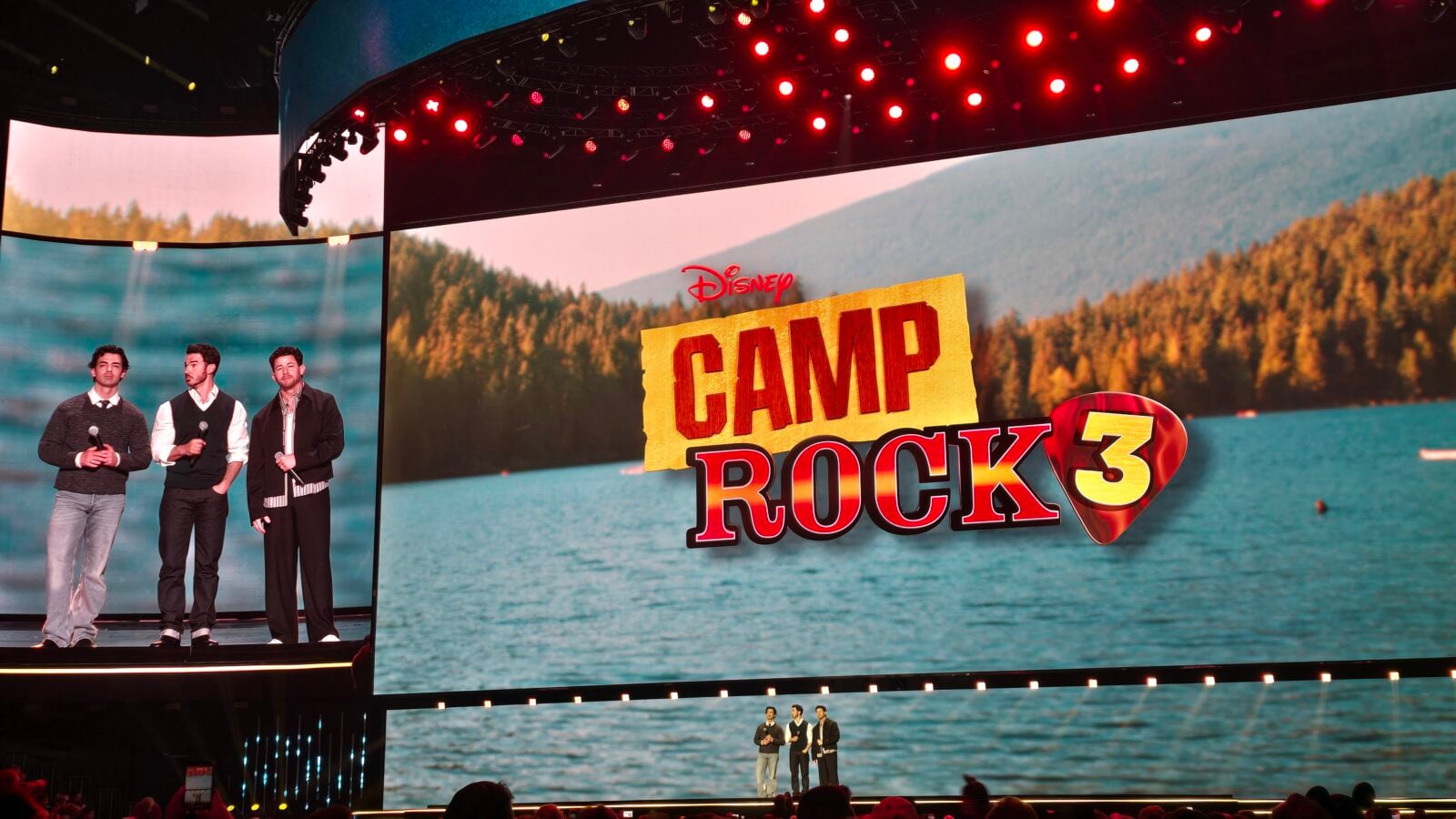 Three male performers with microphones on a stage lake and forest backdrop Disney Camp Rock 3 sign visible on screen behind them