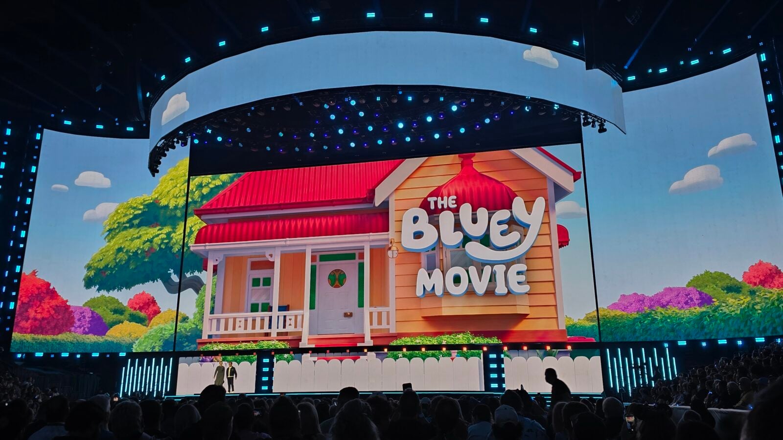 Stage at a live event with a curved screen showing a colorful The Bluey Movie house audience in foreground blue lighting overhead on dark venue