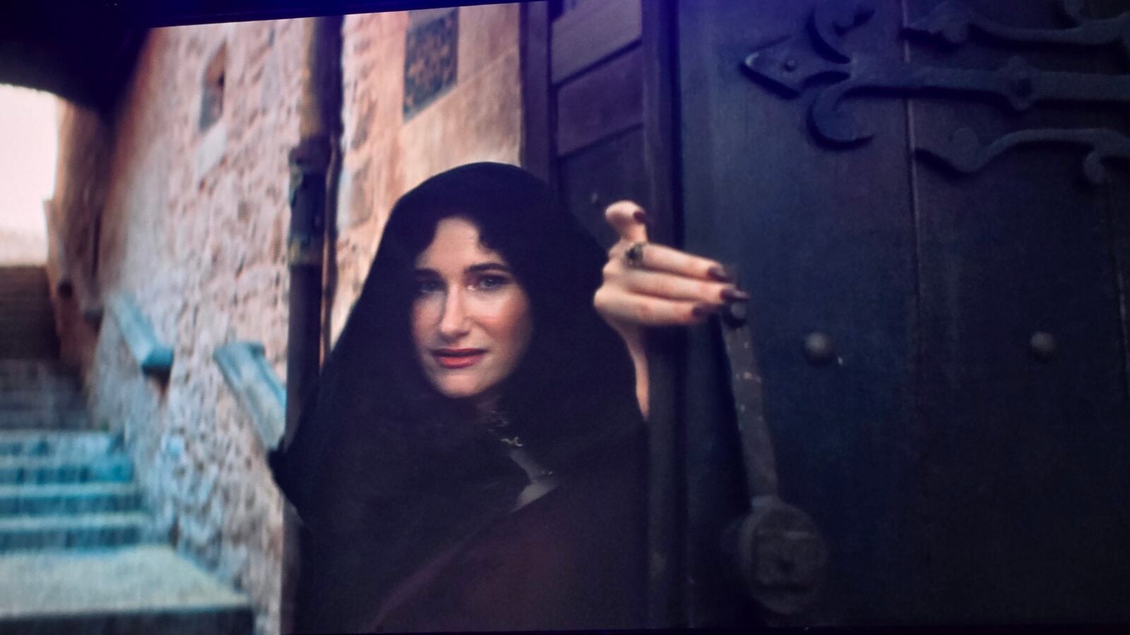 Woman in a black hooded cloak raises her hand in front of a dark gate or door in a moody urban setting