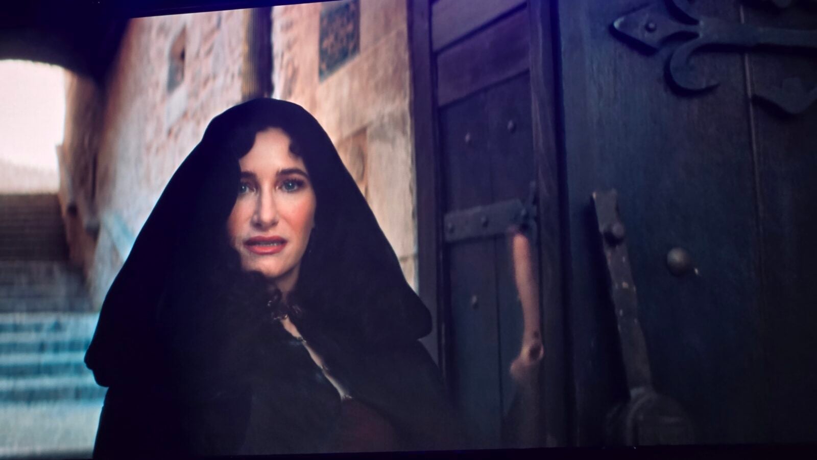 Woman in a black hooded cloak stands beside a dark wooden door facing the camera
