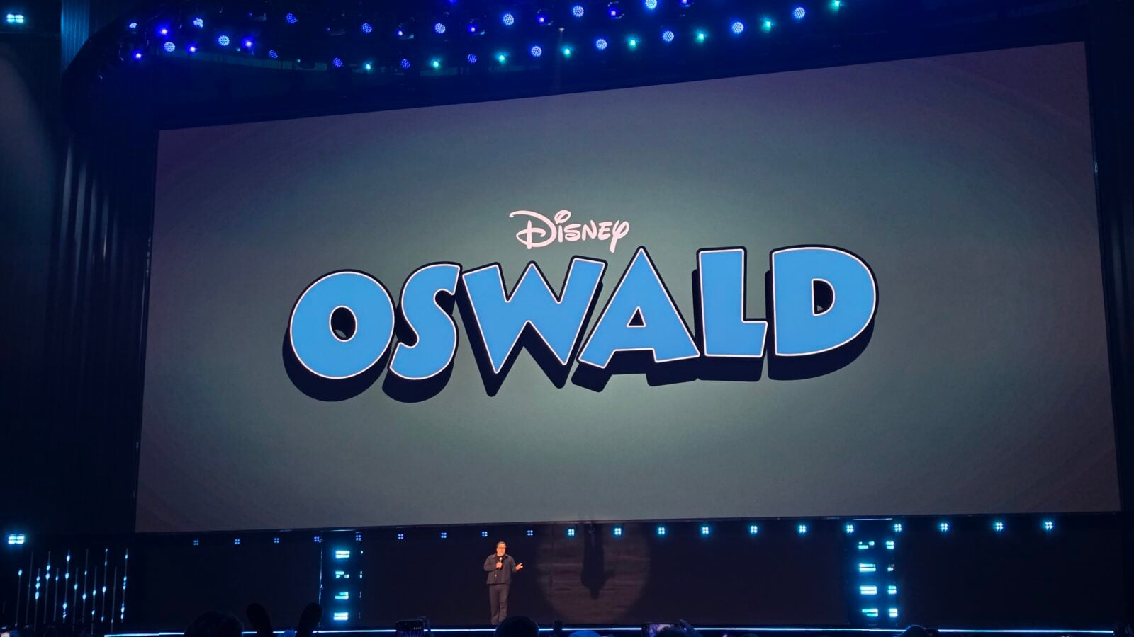 Large stage with a big screen showing Disney Oswald in blue letters a presenter stands at the front with a microphone
