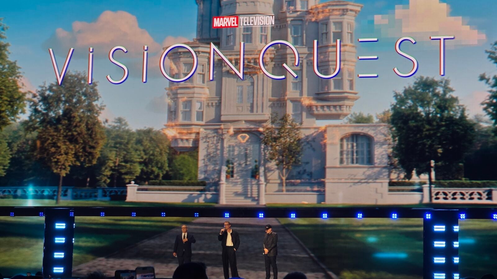 Three men in suits speak on a dark stage with a grand mansion image projected behind them at a Marvel Television event
