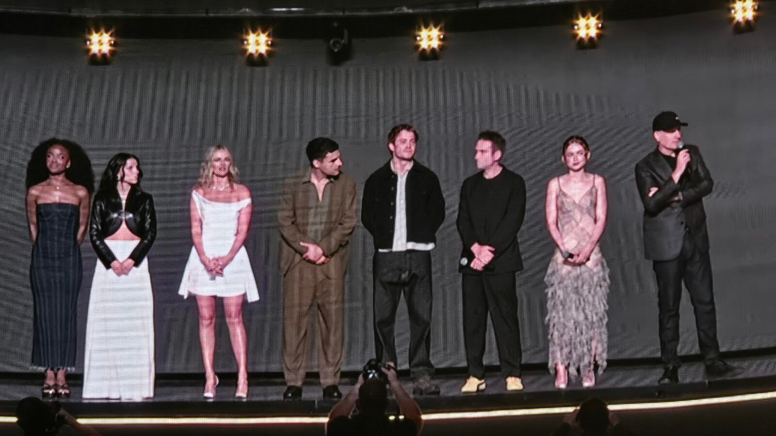 Group of models on a runway stage wearing diverse outfits with a host speaking into a microphone on the right