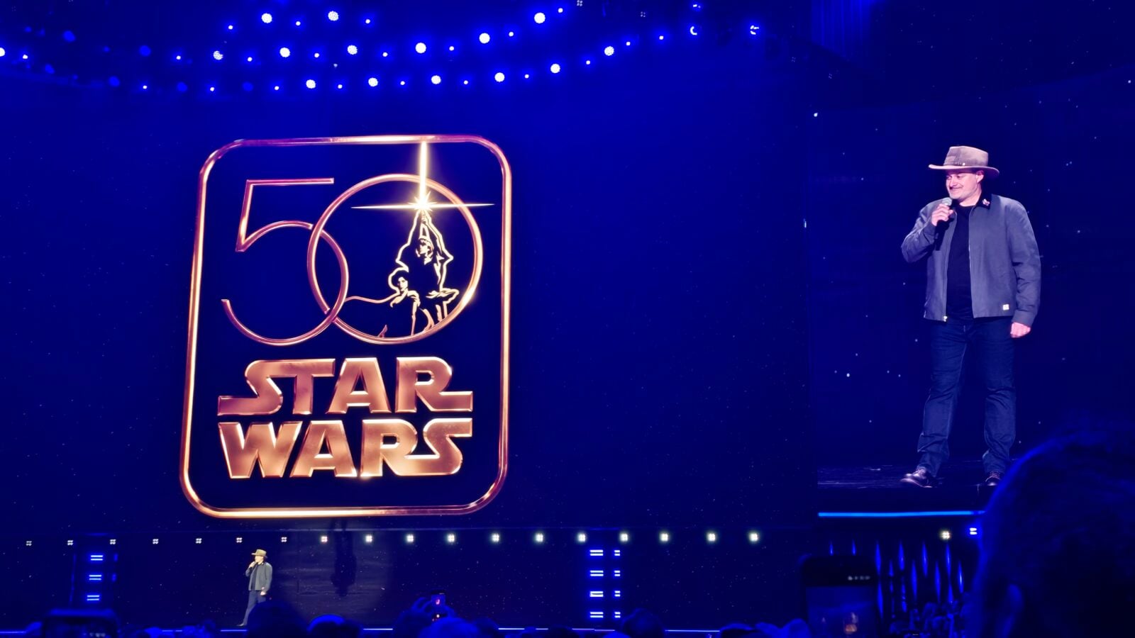 Stage with a large glowing Star Wars logo a man in a hat speaks into a microphone on the right hand side of the stage