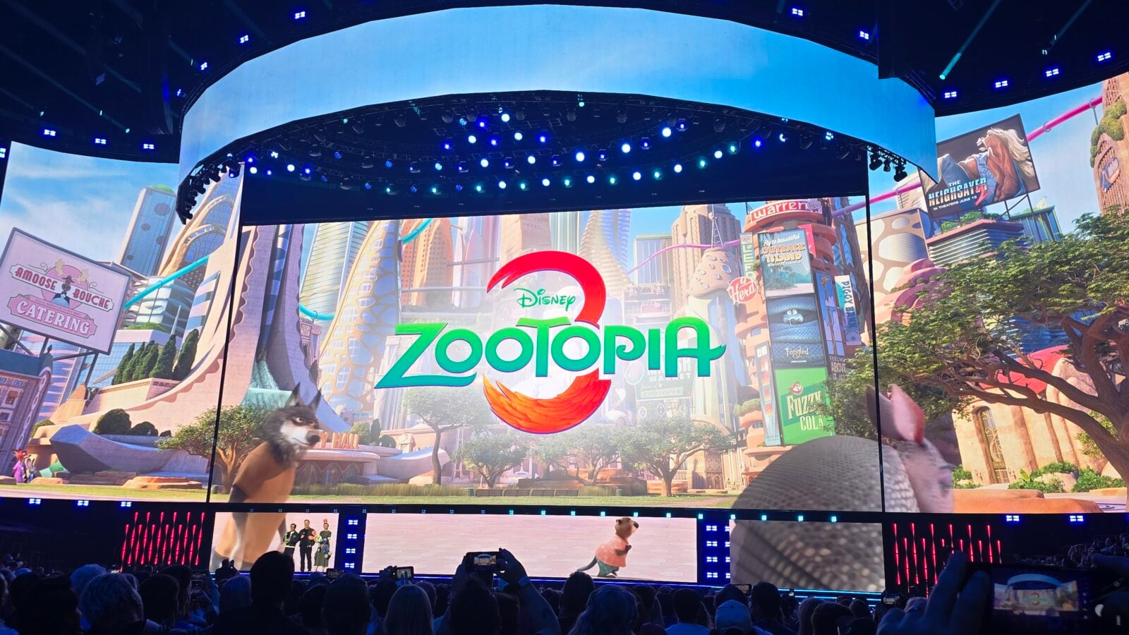 Stage at a convention or show featuring a large screen with the Disney Zootopia logo and colorful cityscape backdrop audience visible in the foreground