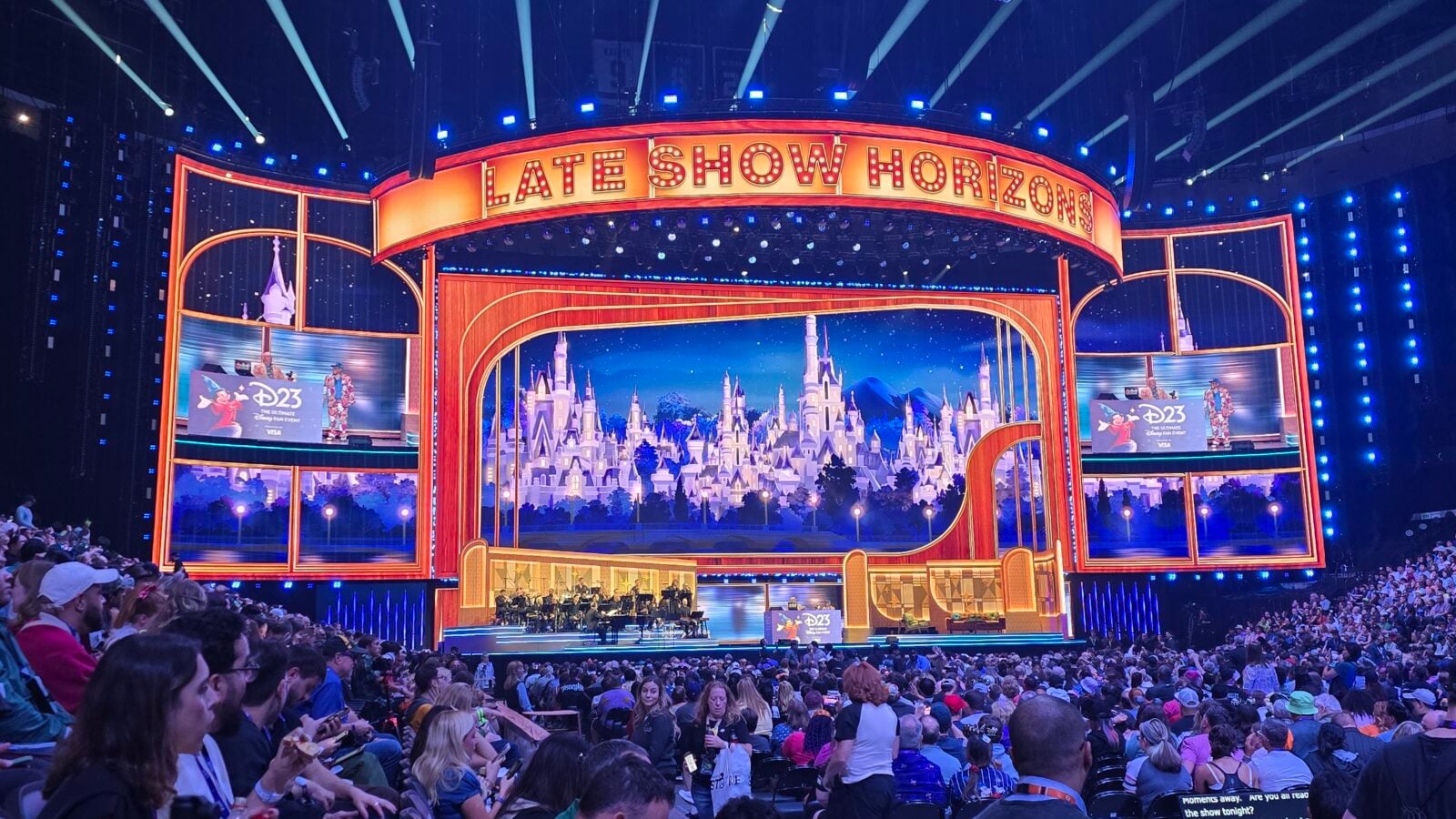 Bright grand theater stage with an orange lit set and a large LATE SHOW HORIZON sign overhead a castle backdrop on stage orchestra members seated below and a full audience watching the performance