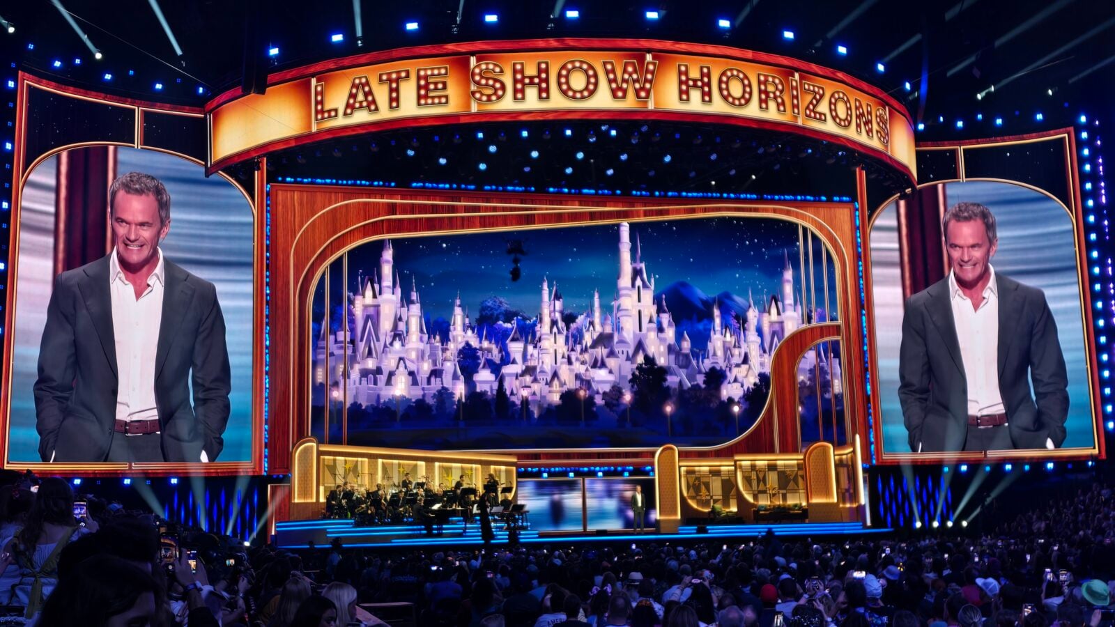 Wide shot of a flashy TV studio stage labeled Late Show Horizons with two side screens showing a man in a suit ornate gold trim and an orchestra performing below the set