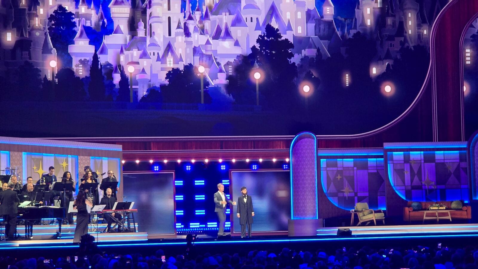 Orchestra performing on a neon lit stage with a castle mural backdrop and two hosts speaking at center