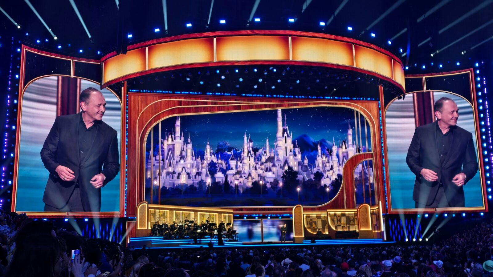 Two men in black suits stand on a bright game show stage with a castle backdrop and an audience watching