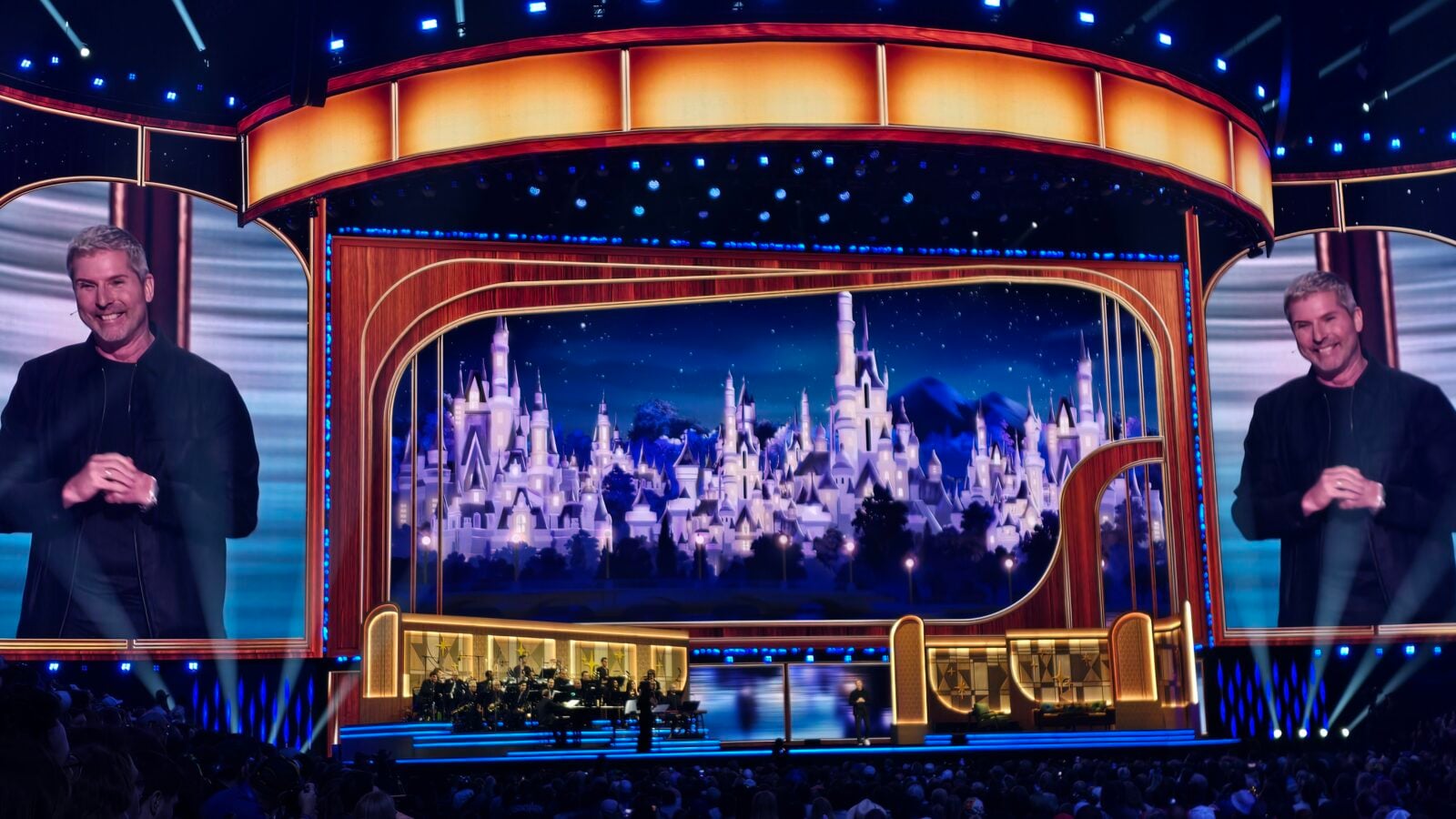 Stage with a curved orange lit backdrop and a castle image on a large screen two men in black jackets stand at each side smiling with an audience below