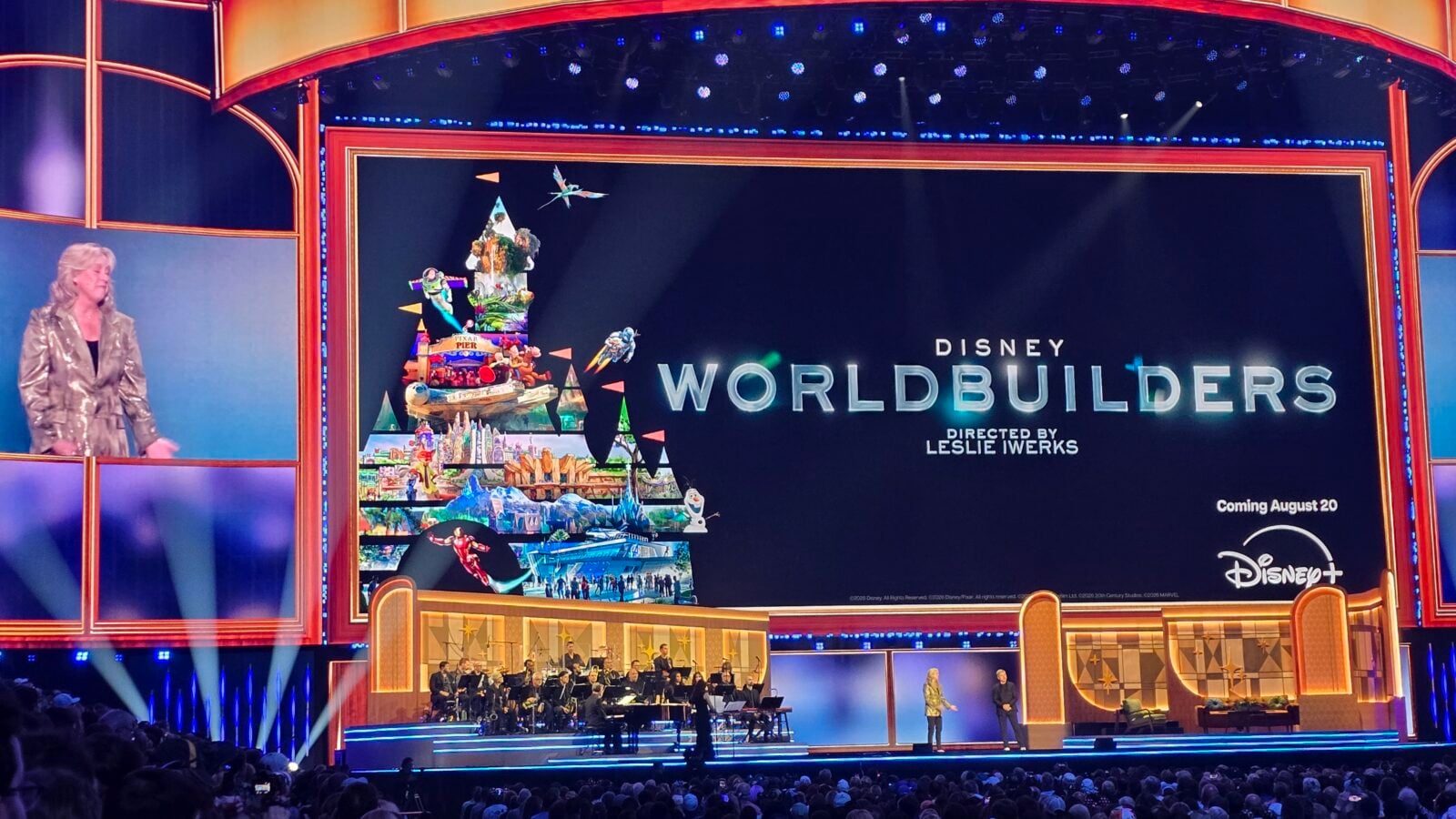 Stage at a Disney event with a large screen reading DISNEY WORLDBUILDERS and an orchestra performing below