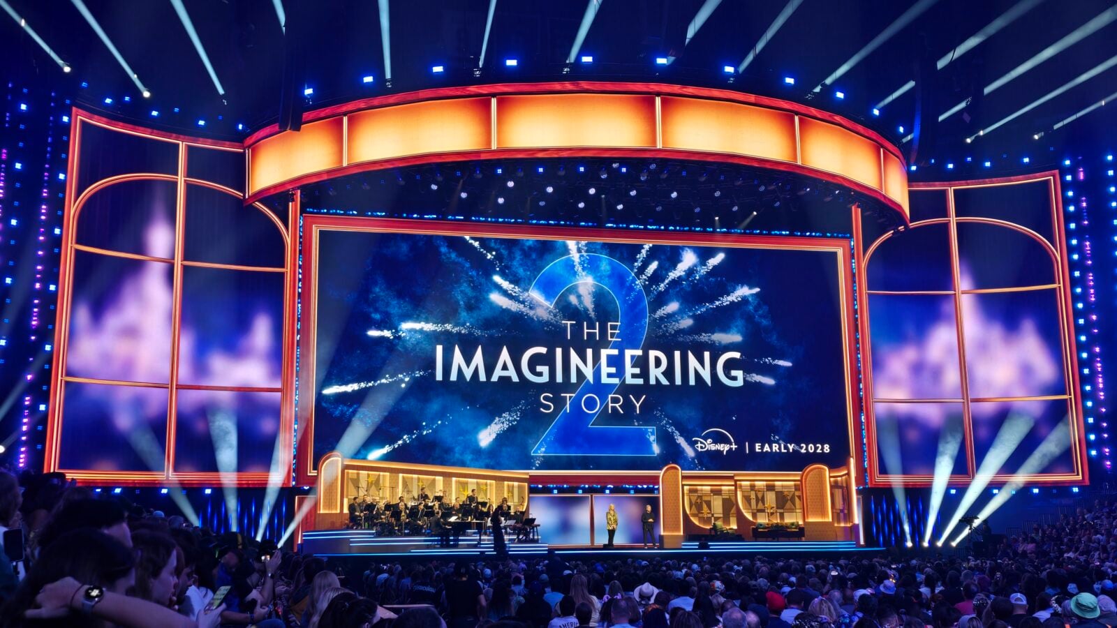 Large theater stage with blue and orange lighting a curved orange surround and a central screen reading THE IMAGINEERING STORY as an audience watches