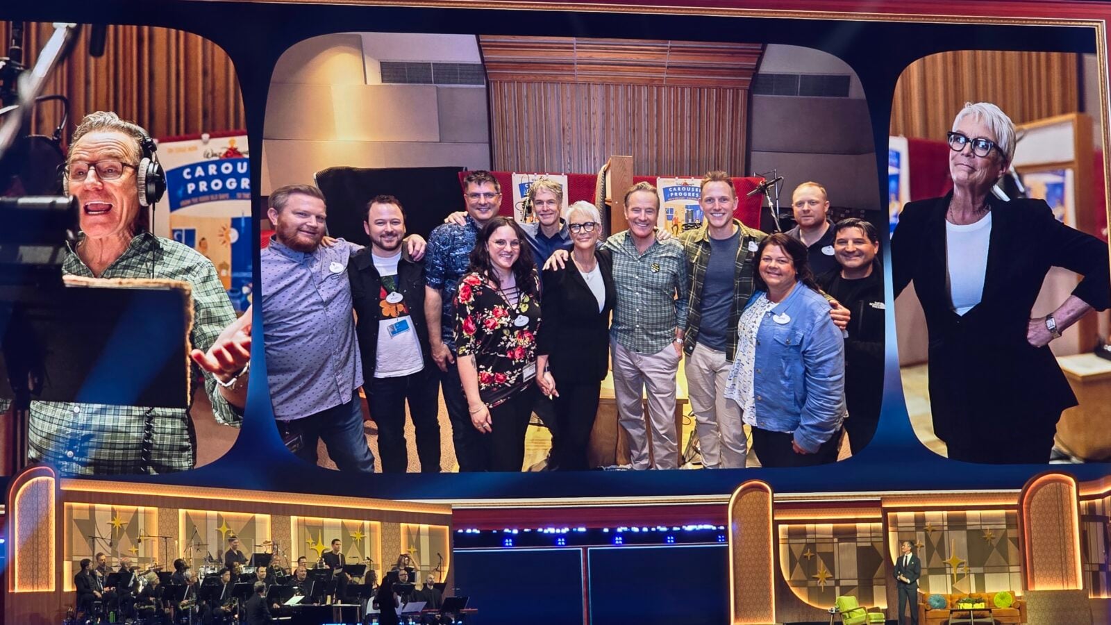 Group of about a dozen people posing for a photo on a TV shaped stage backdrop with a live band visible in the lower left corner