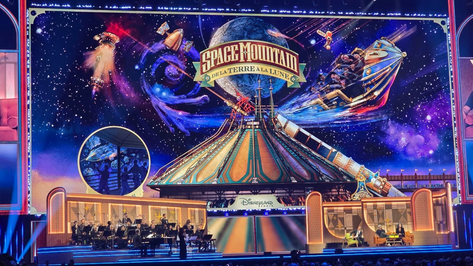 Broadway style stage with a colorful Space Mountain backdrop and an orchestra performing in front