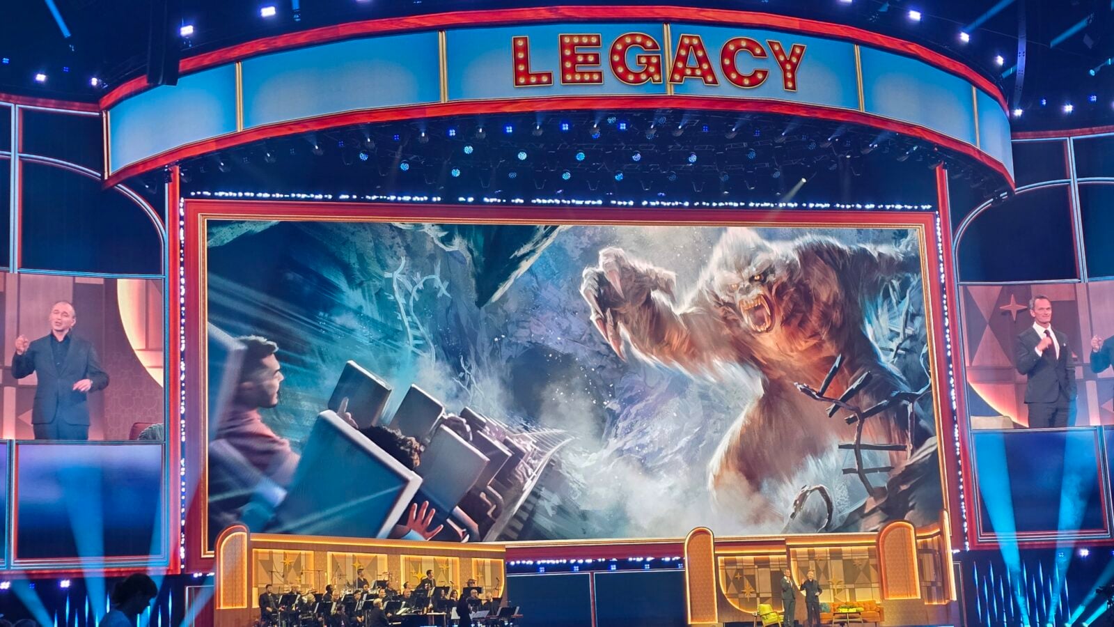 Colorful game show stage with a red blue marquee reading LEGACY a large central screen showing action two hosts at the sides and an orchestra in front
