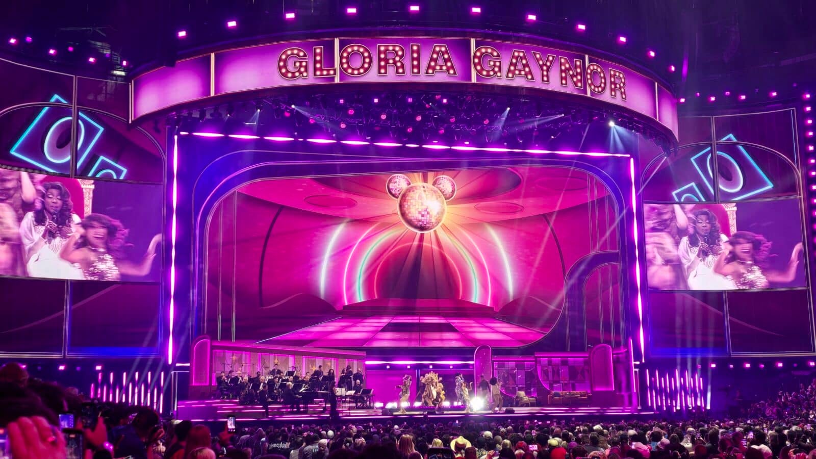 Gloria Gaynor concert stage with purple lighting a large sign disco ball centerpiece and an orchestra performing