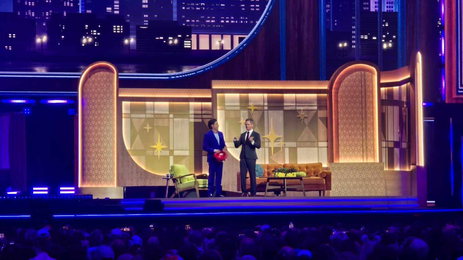Two men in suits stand on a brightly lit retrothemed TV stage conversing as a live audience watches from below