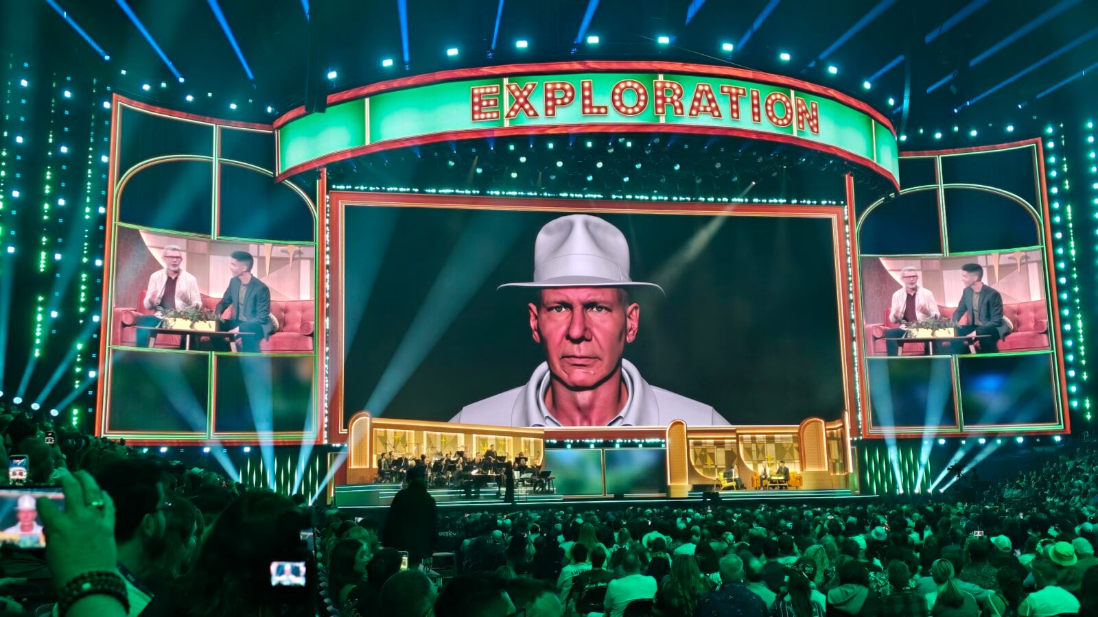 Neon lit stage with a large central screen showing a man in a light fedora audience filming with phones in the foreground