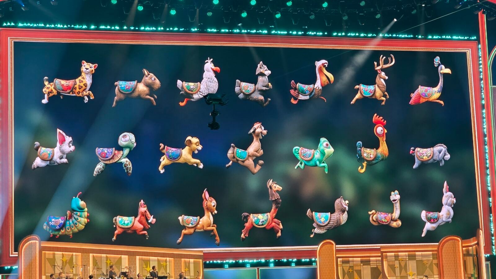 Colorful row of whimsical animal puppets horses birds turtle suspended across a theater stage with bright turquoise lights and a red gold frame behind