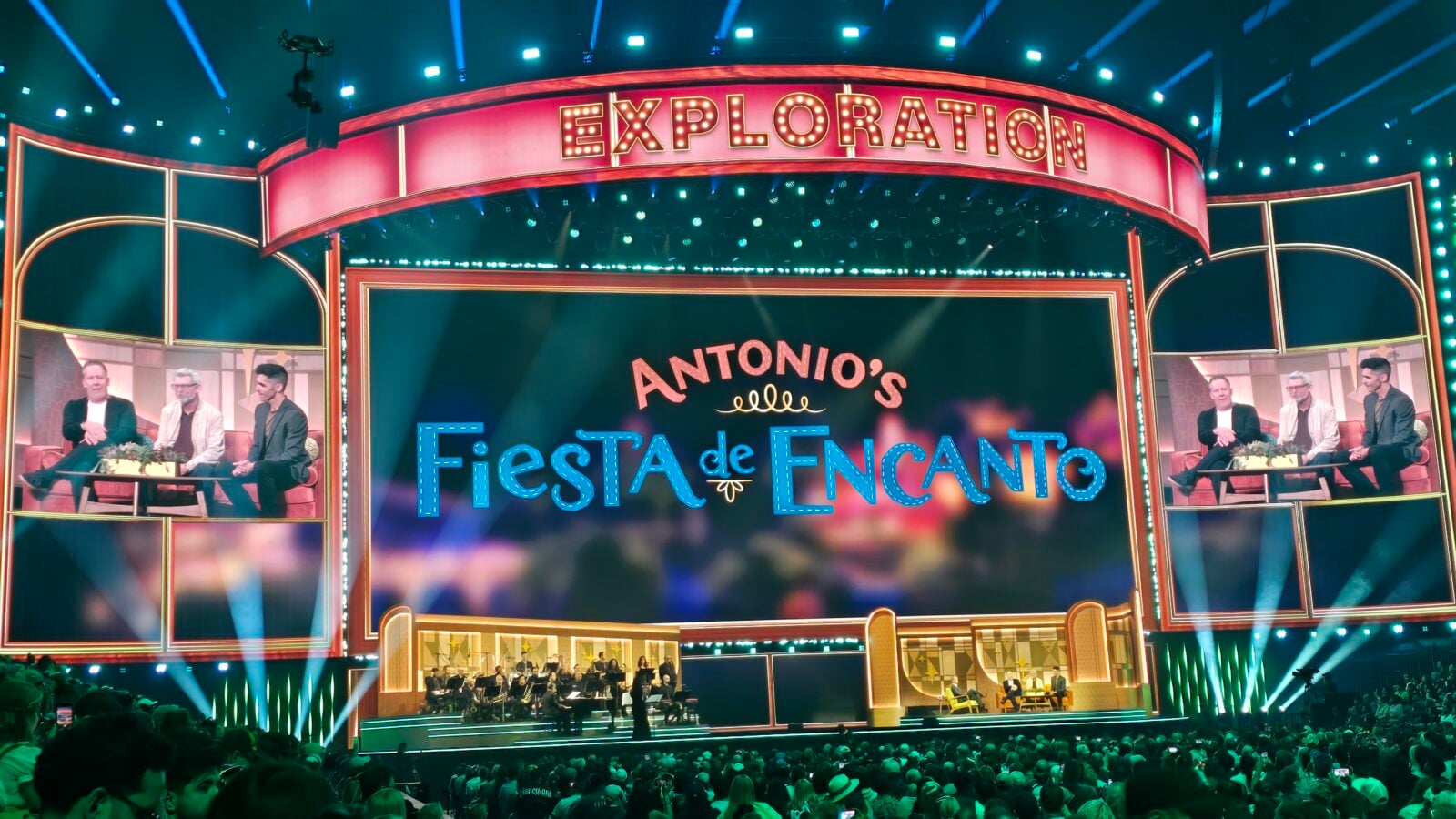 Vibrant stage set with a red marquee reading EXPLORATION a central screen showing Antonios Fiesta de Encanto and a panel of speakers on each side an orchestra performs below with a cheering audience in the foreground