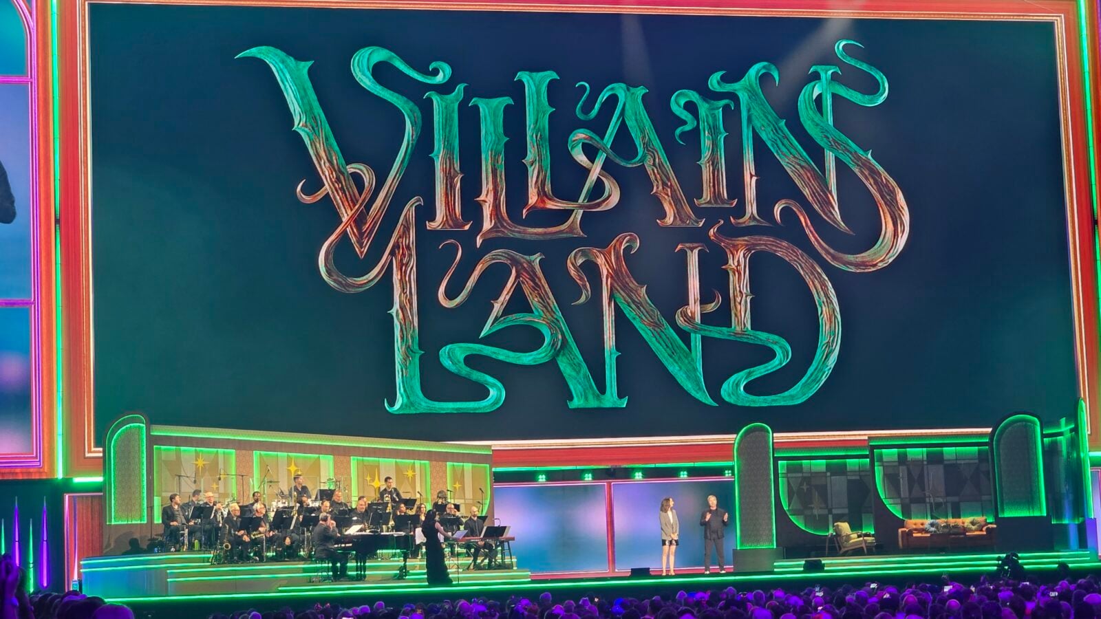 Orchestra performing on a neon lit Las Vegas stage with two presenters and a large decorative Viva Las Vegas backdrop above
