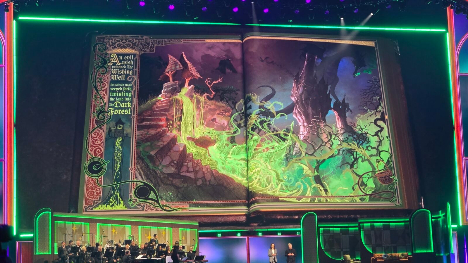 Stage shows a giant open book prop with neon green borders an orchestra seated below and two presenters at the front