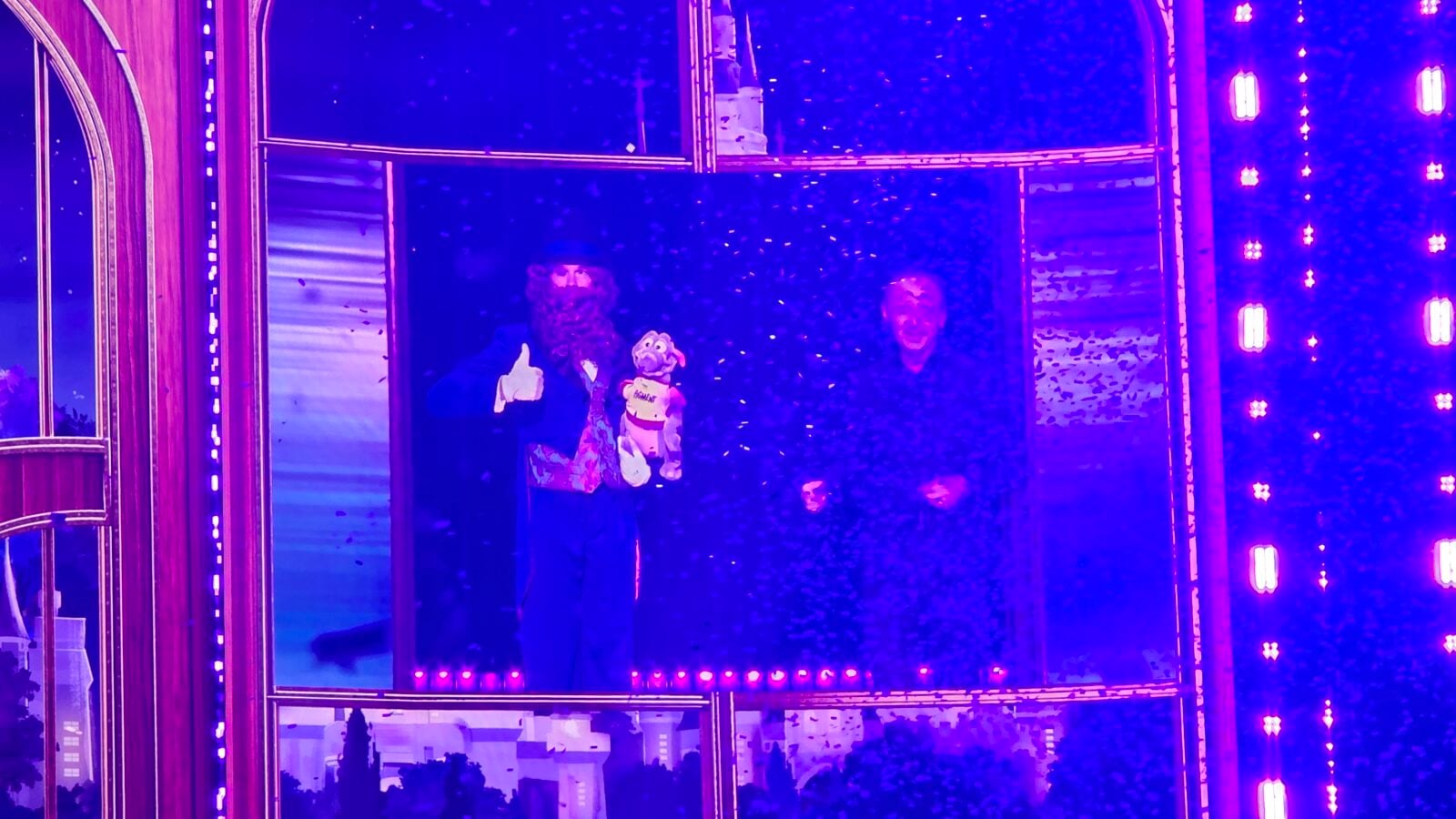 Two men in a glass enclosed stage box under purple lights one gives a thumbs up while the other holds a stuffed toy