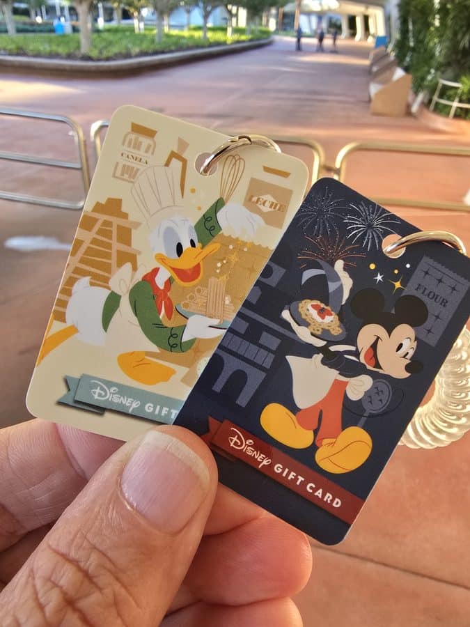 Two Disney gift cards held between fingers one with Donald Duck artwork and the other with Mickey Mouse foreground focus with a blurred walkway in the background