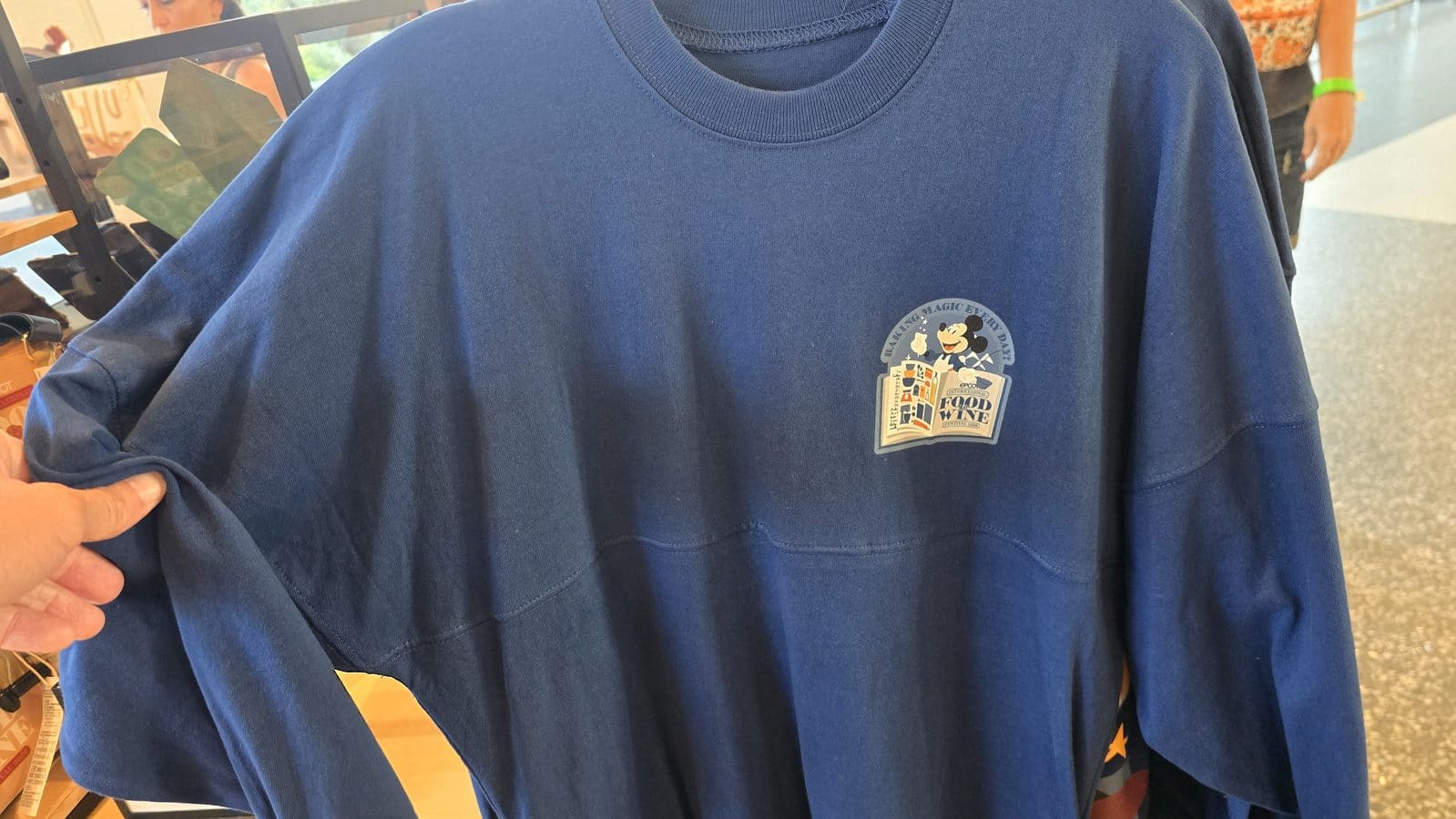 Blue sweatshirt with a small graphic of Mickey Mouse reading a magazine featuring the text FOOD  WINE on the badge