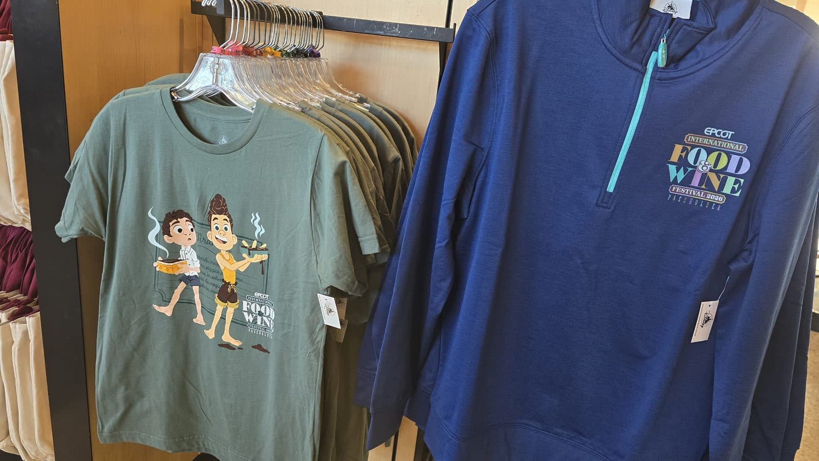 Green t shirt featuring cartoon characters holding food hanging on a rack beside a blue zip up jacket with a colorful Food  Wine Festival 2026 logo