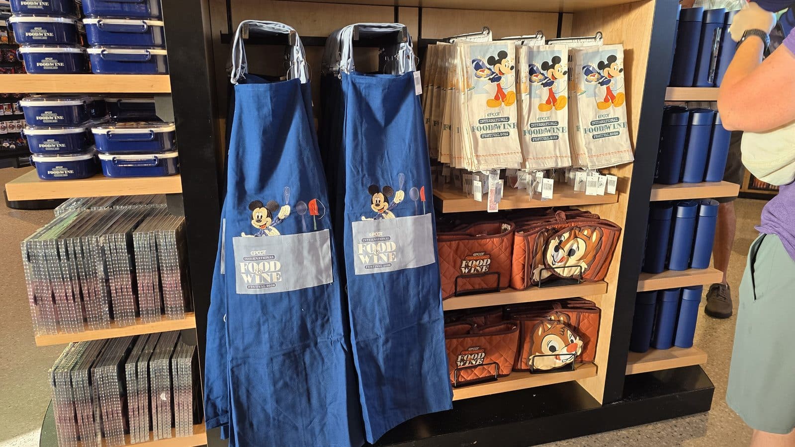 Display of blue aprons and beige tote bags with Mickey Mouse and Food  Wine Festival logos on a shop shelf stacks of blue containers to the left and other merchandise around