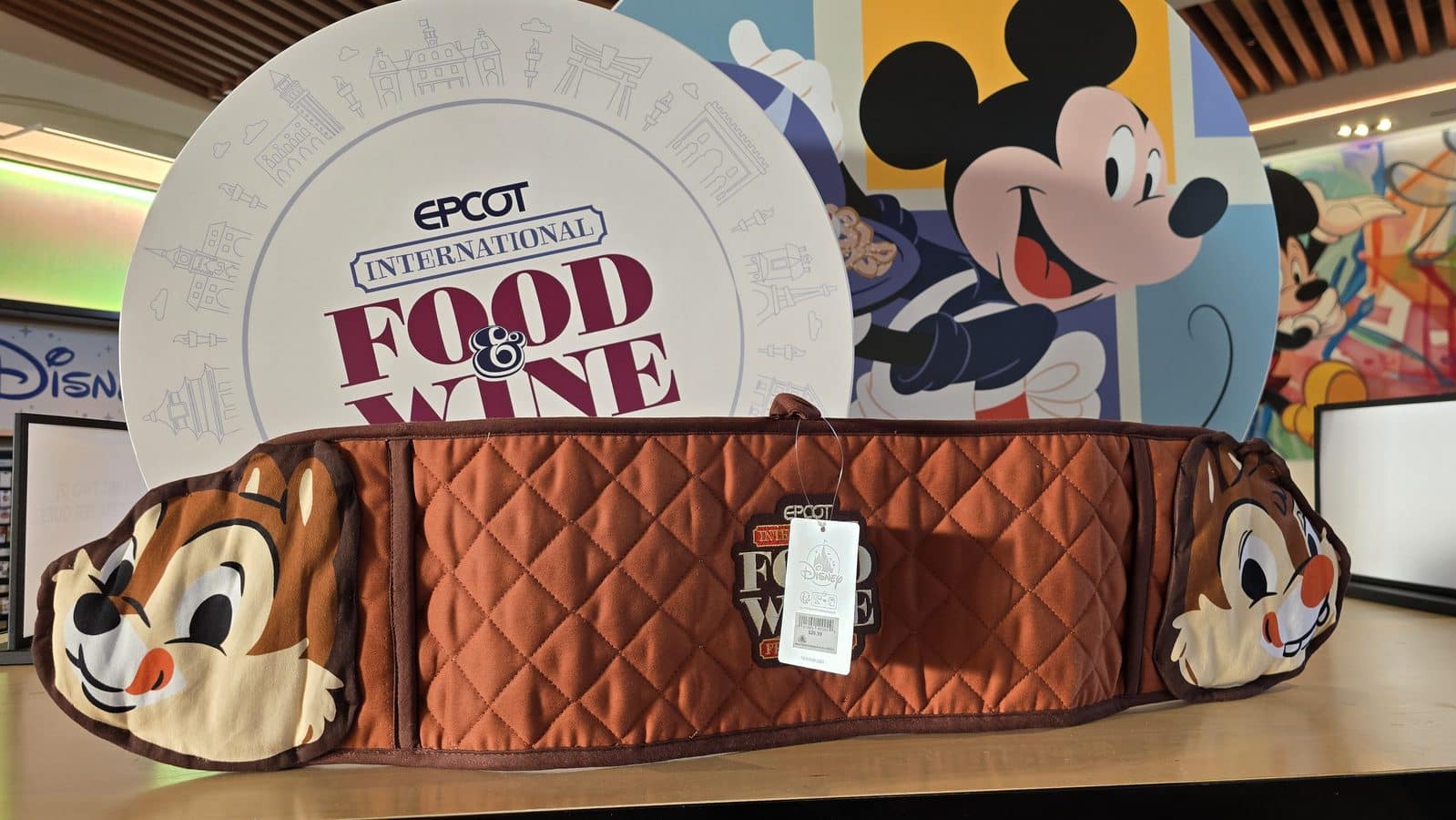 Brown quilted Disney themed bag in the foreground with a large round display reading Epcot International Food  Wine and a colorful Mickey Mouse mural in the background