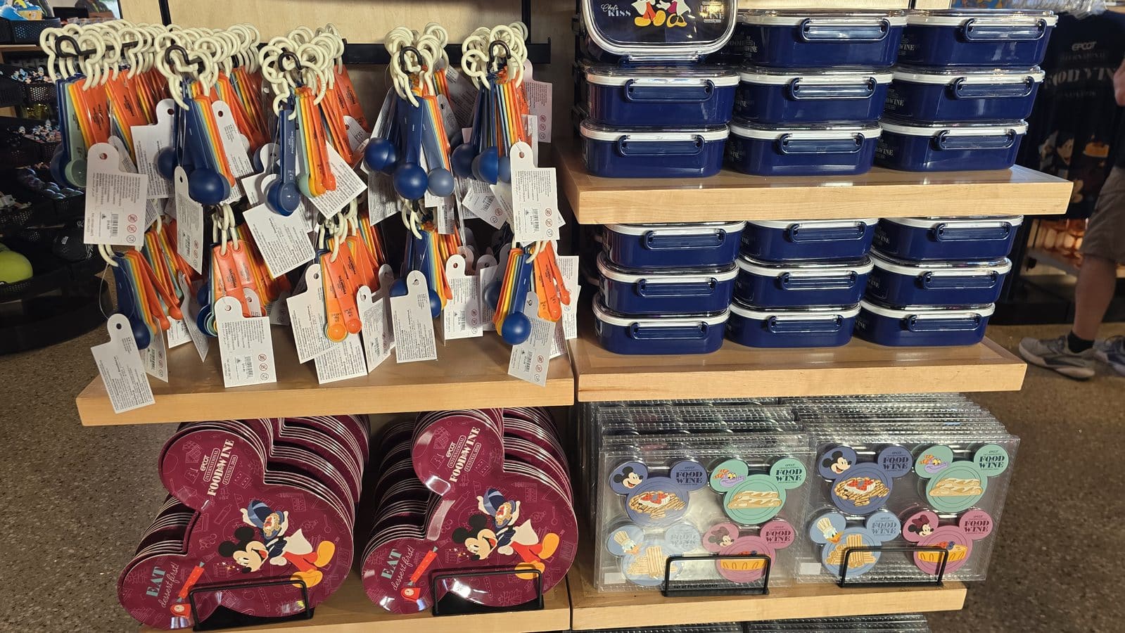 Row of hanging colorful measuring spoons and keychain tags on the left with stacked blue lunch boxes on shelves to the right