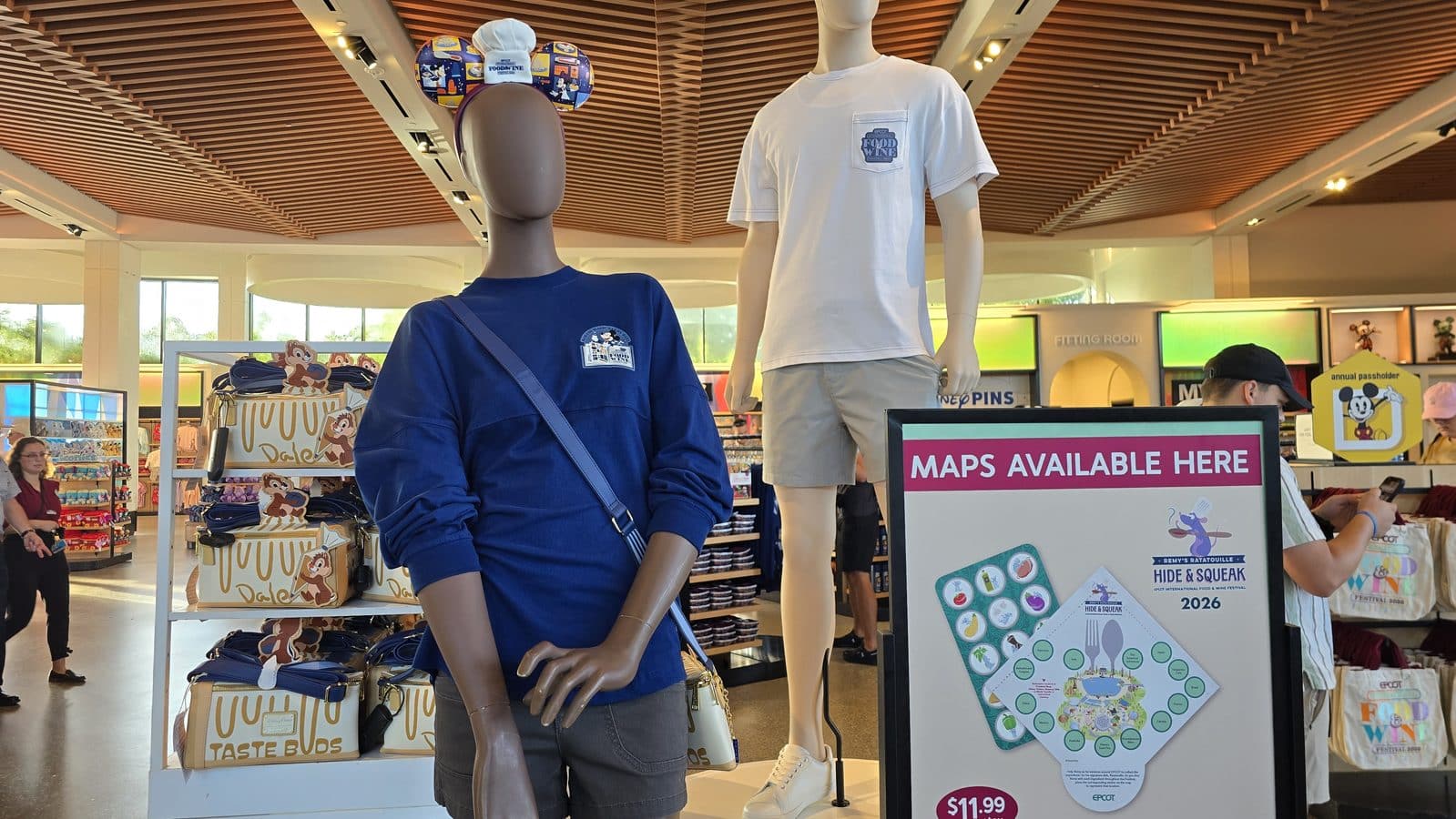 Two mannequins wearing blue and white t shirts in a bright store display with merchandise racks in the background and a Maps Available Here sign nearby